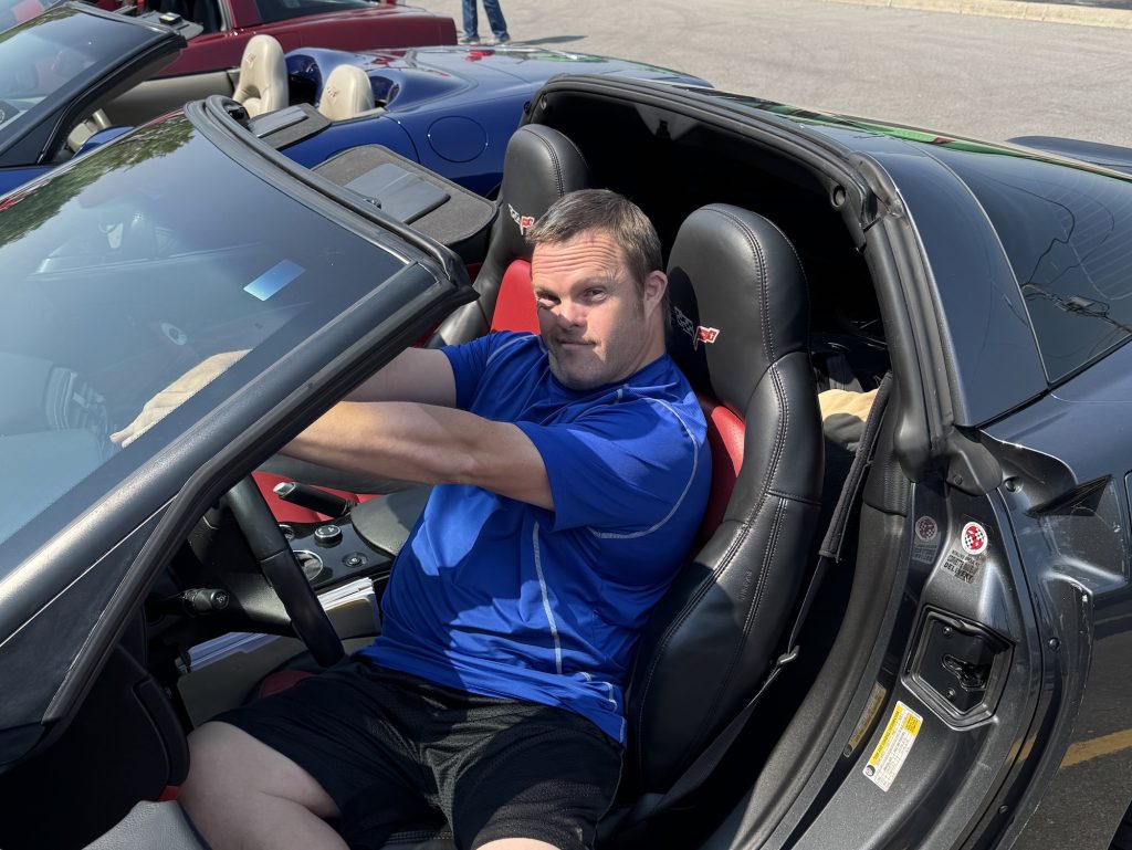 Chris W suavely poses in the driver seat of a cool convertible car.