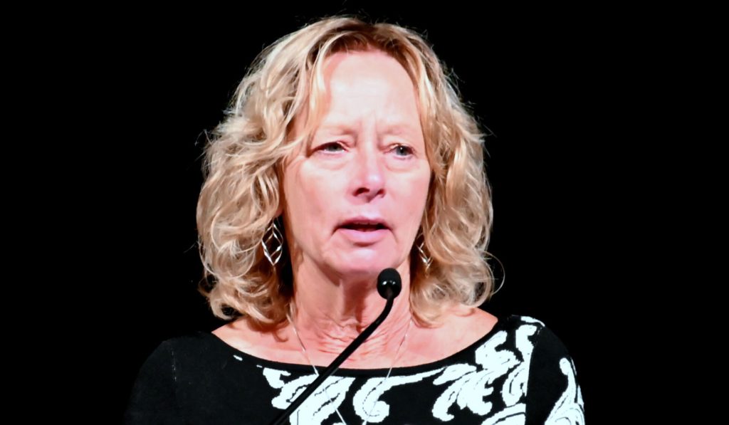 Susan Mielcarek wearing a black and white shirt. She stands at a podium with a microphone near her chin.