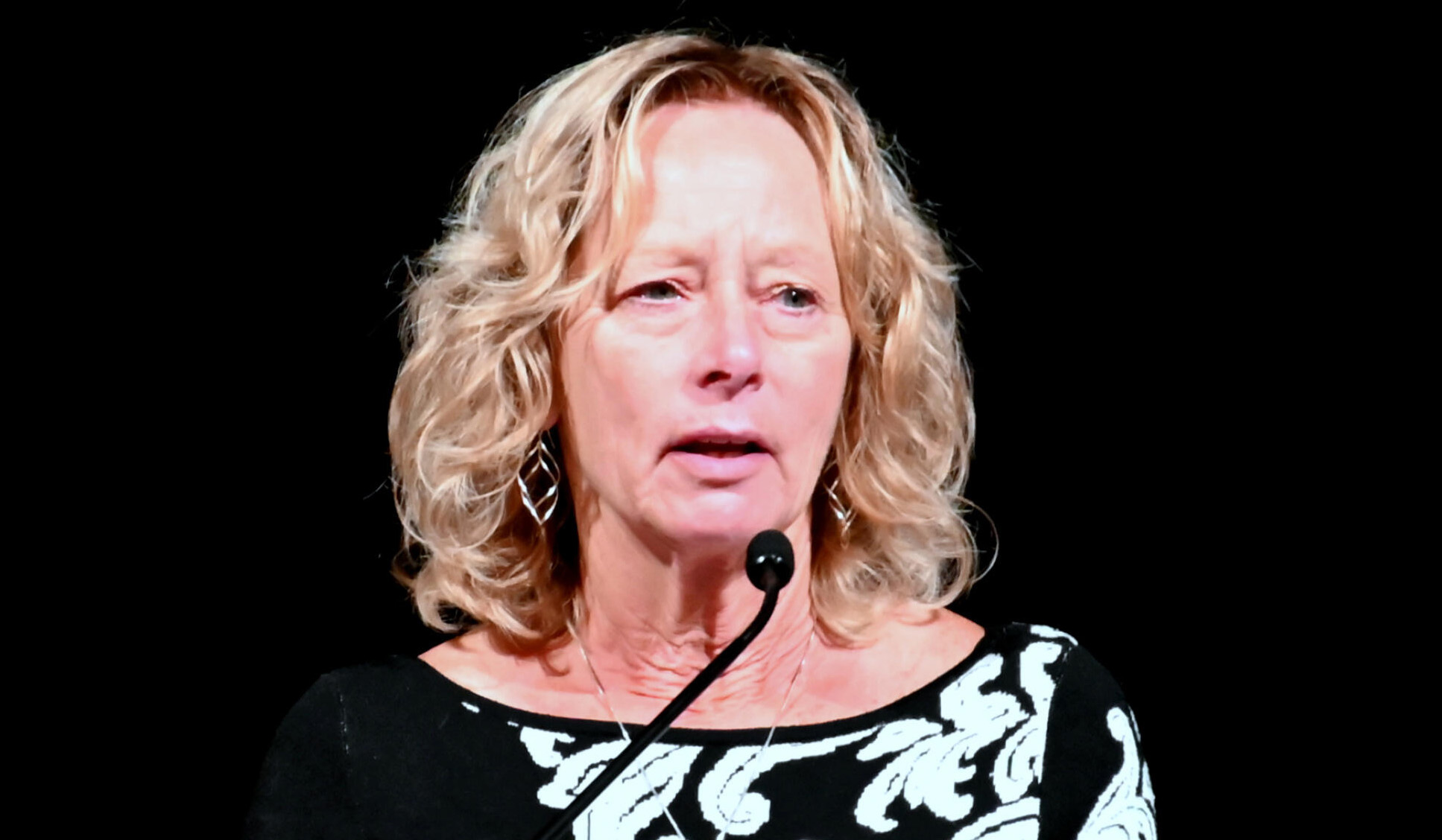 Susan Mielcarek wearing a black and white shirt. She stands at a podium with a microphone near her chin.