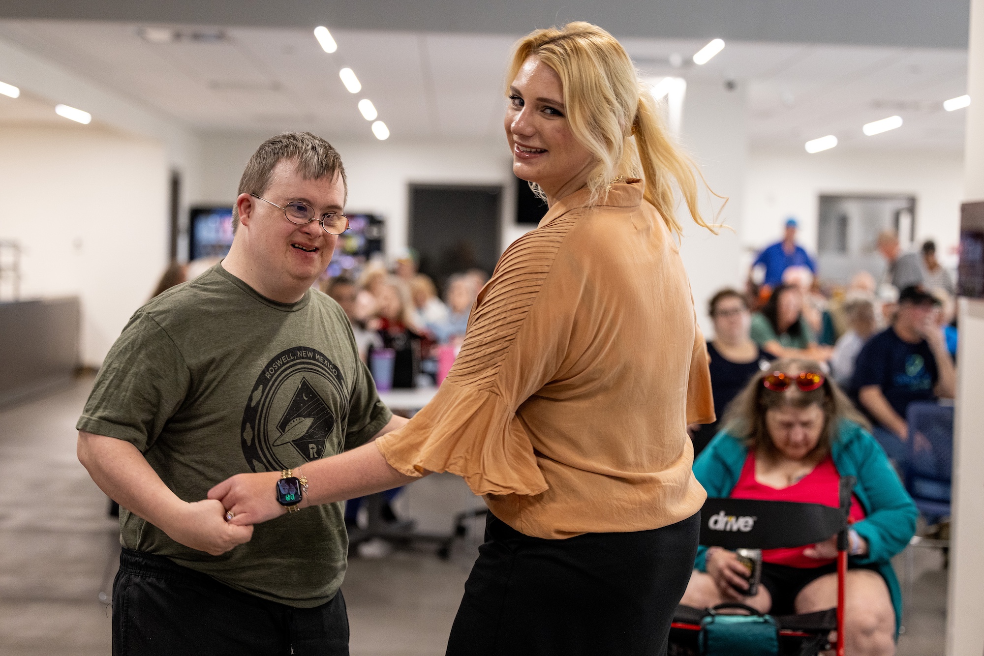 A woman in a neutral colored shirt looks over her shoulder at the camera while she dances with a young man in glasses and a green tee shirt.