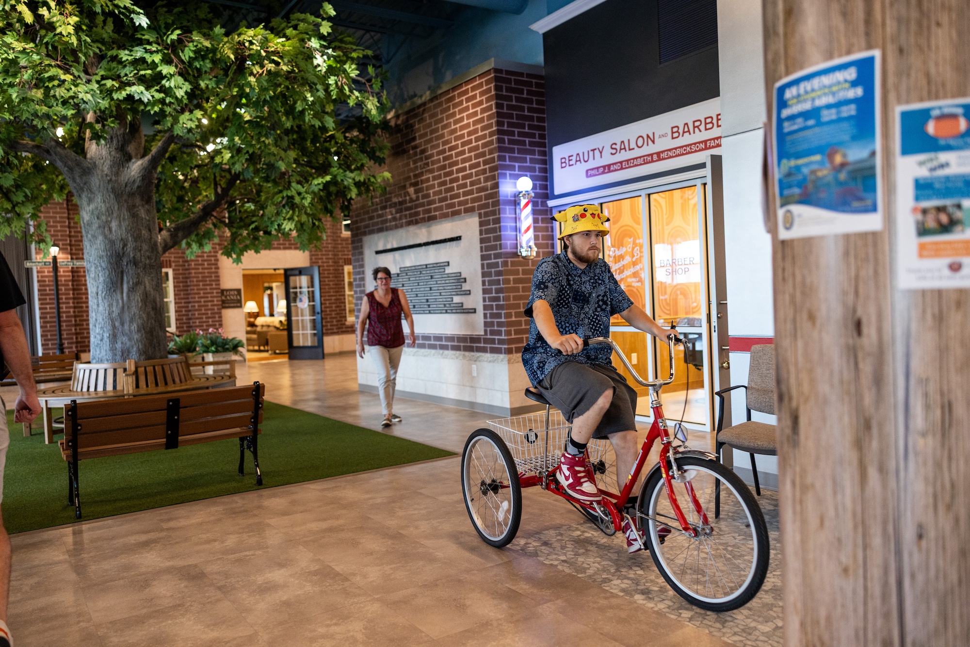 A man in a bright yellow PIkachu-themed bucket hat rides an adult-sized tricycle through Yesteryear Village.