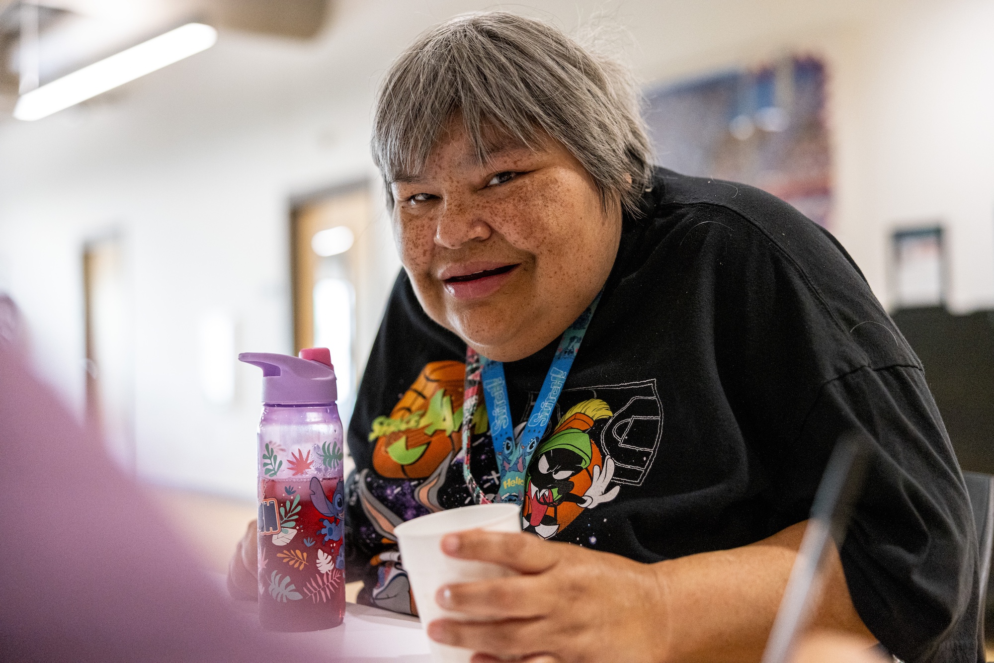 A woman wearing a Space Jam tee shirt leans over a table and looks at the camera.