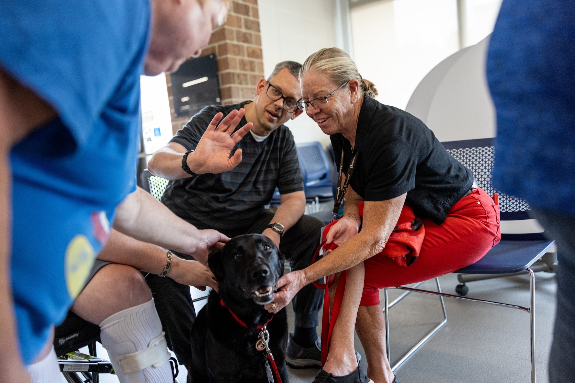 A man waves to the camera while the woman beside him scritches the chin of a happy black lab dog.