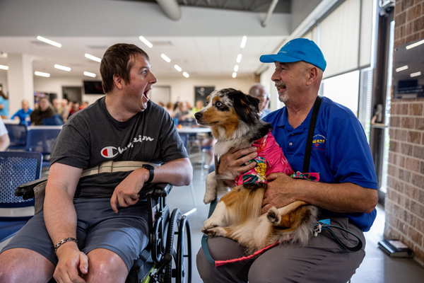 A man in a wheelchair looks very excitedly at a man smiling and sitting next to him holding a dog on his lap