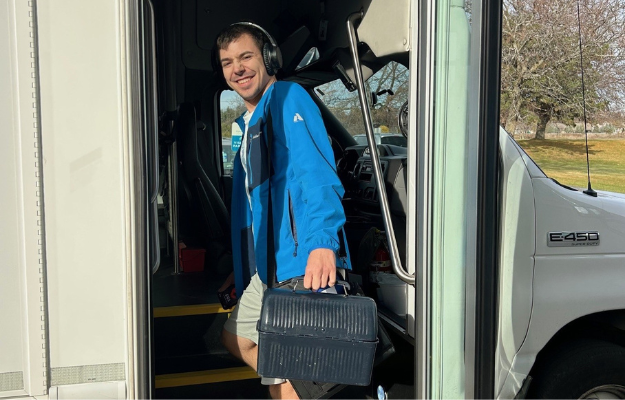 A man wearing headphones and holding a small plastic bin with a handle is getting on a bus