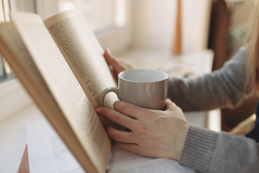 A woman reading a book and drinking out of a mug