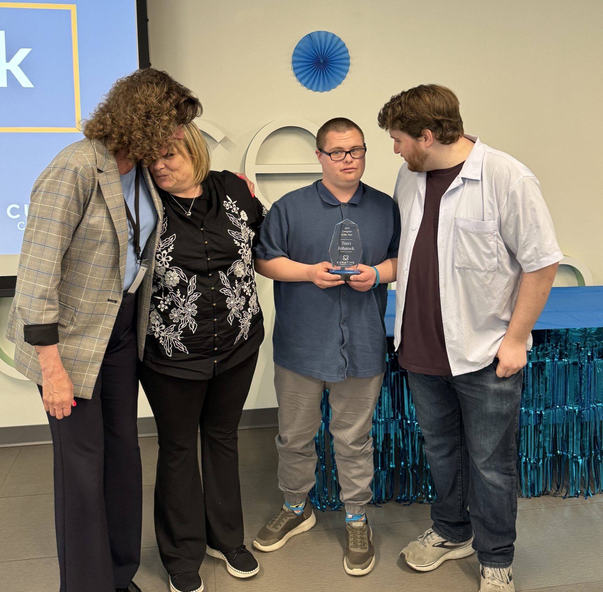A line of four people getting ready to pose for the camera in front of a projector screen. A man in a blue shirt holds a glass award in his hands.