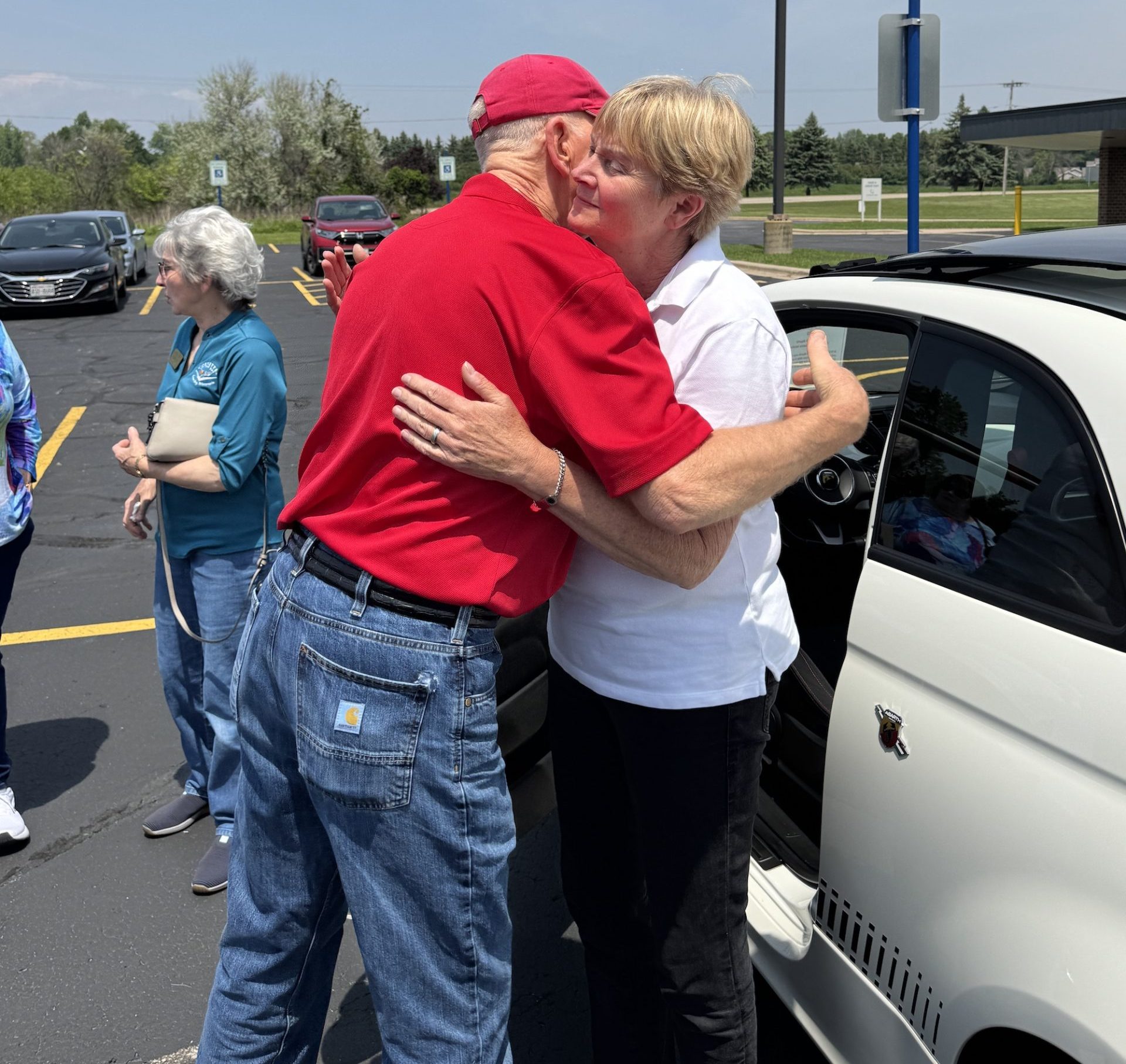 A woman in a white shirt and a man in a red shirt and baseball cap share a hug outside in a parking lot next to a white car.