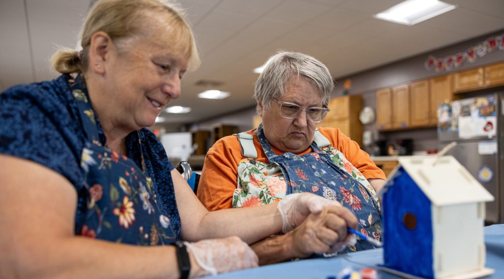 Two women wearing blue floral aprons concentrate as they paint a birdhouse blue.